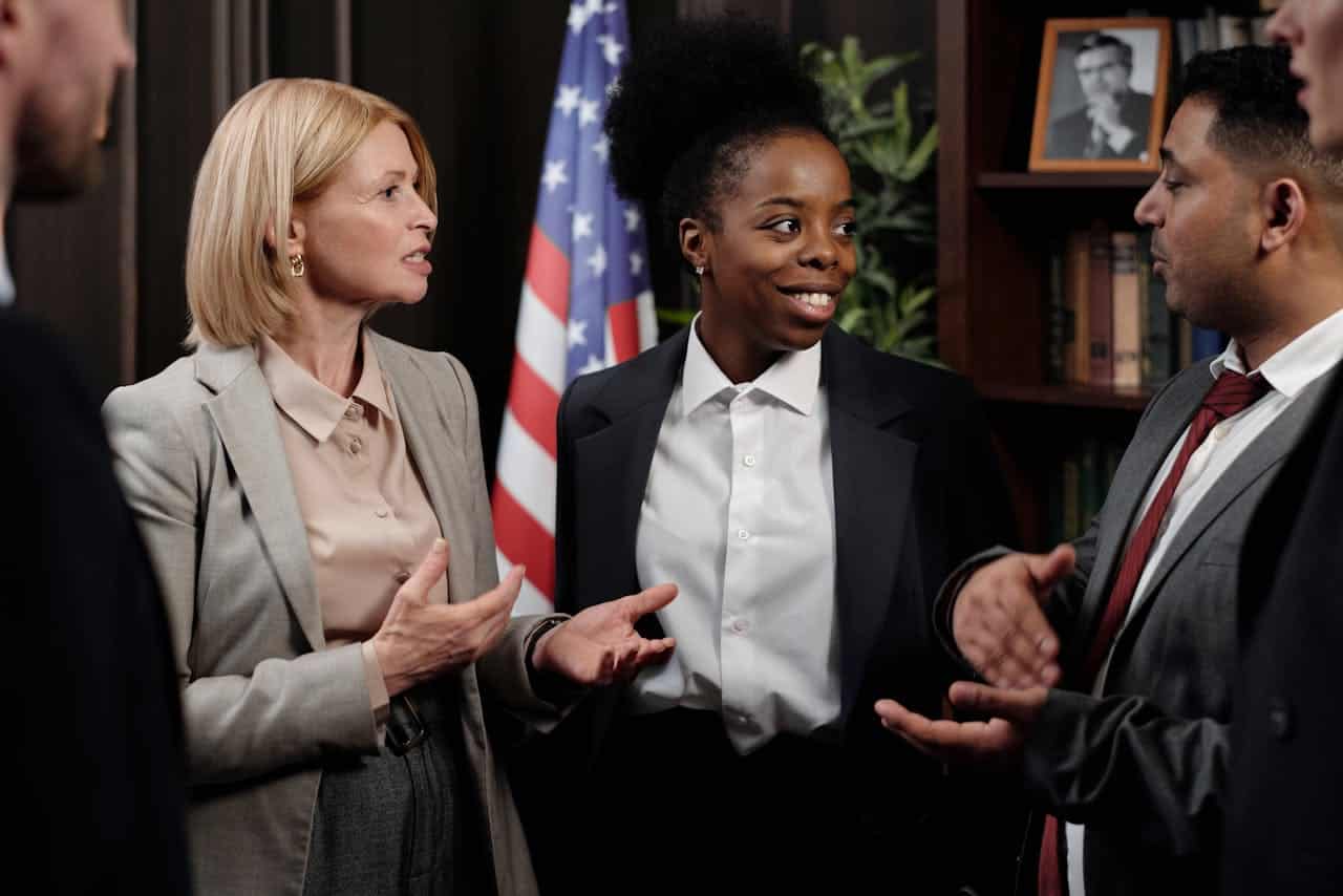 May contain four people in business attire, with an American flag in the background.