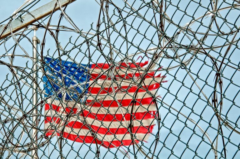 May contain an American flag behind a chain-link fence and razor wire.
