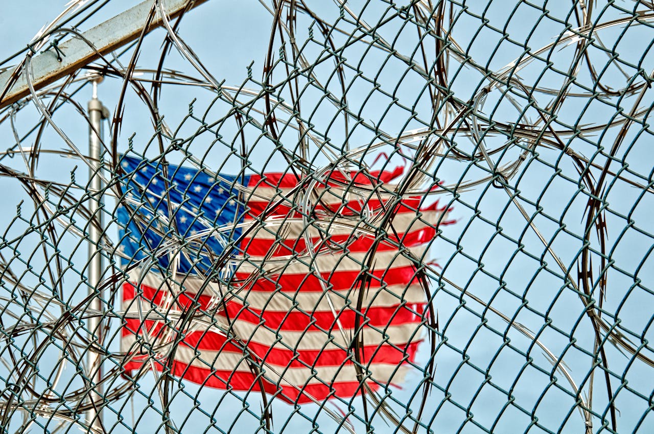 May contain an American flag behind a chain-link fence and razor wire.