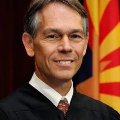 May contain a person in a black robe and a tie, with a flag in the background.