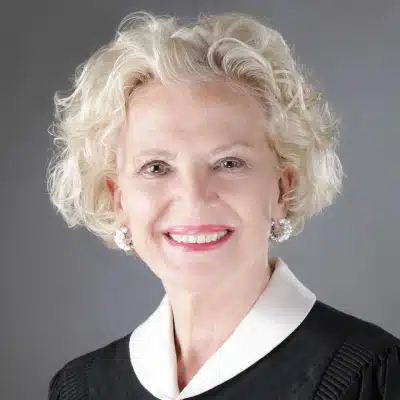May contain a person with short blonde curly hair wearing a black robe and white collar.