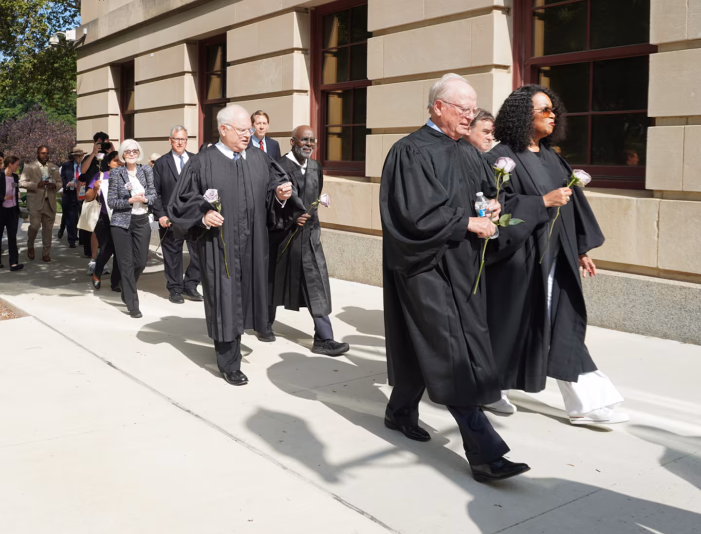 May contain several people in black robes walking outside, holding flowers.