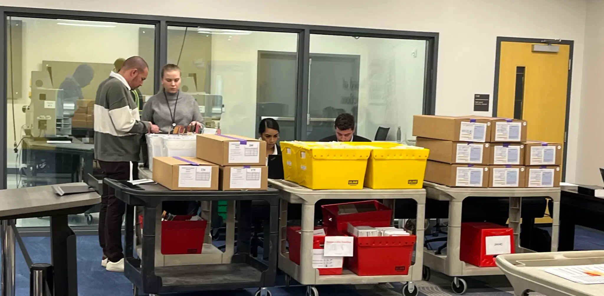May contain three people working at desks with boxes and bins.