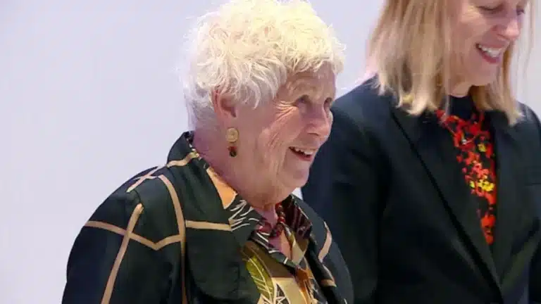 May contain two people smiling. One person has white hair and is wearing a patterned jacket.