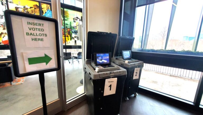 May contain two ballot tabulators with signs and a sign that says "INSERT VOTED BALLOTS HERE" with an arrow.