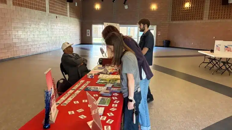 May contain three people looking at a table with pamphlets and stickers.