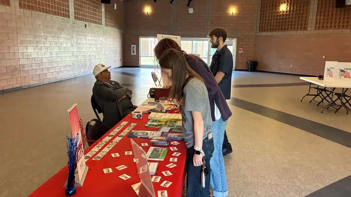 May contain three people looking at a table with pamphlets and stickers.